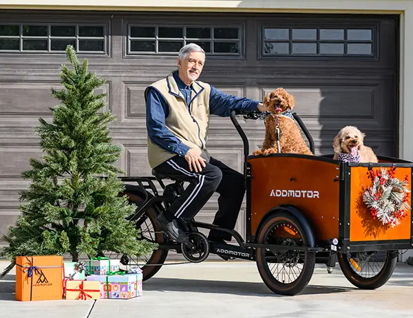 An older man sitting on a two-front-wheel electric trike with two dogs in the front cargo box, surrounded by holiday decorations.