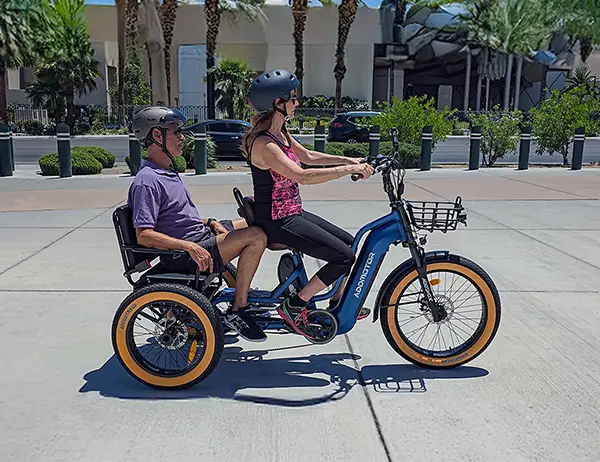 A woman riding an electric trike with a passenger seat, carrying an older adult comfortably on a sunny street.